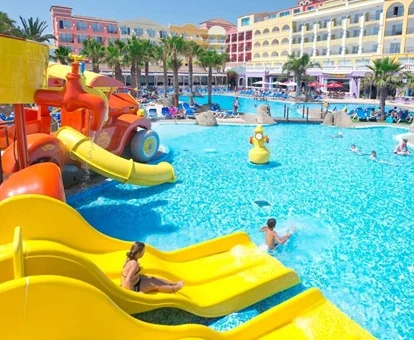 Pasarela de madera entre las piscinas con el bar al fondo y un grupo de adultos tomando una copa