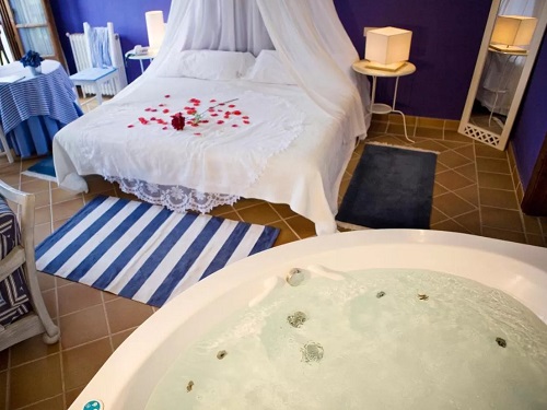 Jacuzzi circular frente a la cama con pétalos de rosas y una rosa sobre las sábanas en la Habitación con cama extragrande y bañera de hidromasaje del hotel rural Llosa de Fombona