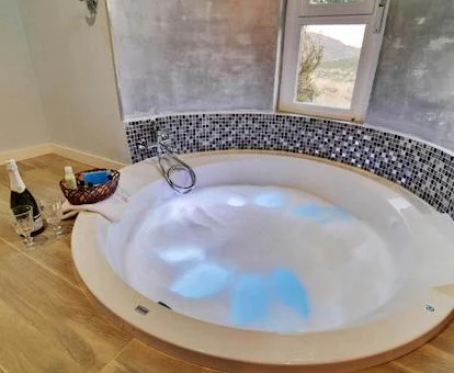Foto del jacuzzi de forma circular que se encuentra a ras de suelo junto a la ventana con vistas al campo y que tiene una botella de Champagne y dos copas junto al borde.
