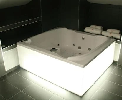 Foto del jacuzzi para dos personas con chorros de hidroterapia en un rincon frente a la cama iluminado por los laterales