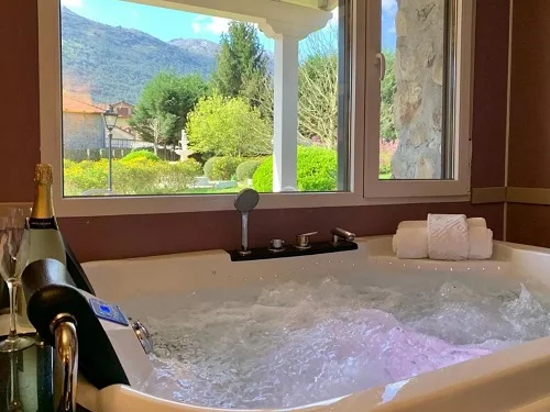 Foto de la Suite con bañera de hidromasaje donde vemos la cama decorada con petalos formando un corazón junto con una caja de bombones y el jacuzzi junto a la ventana de la buhardilla de madera.