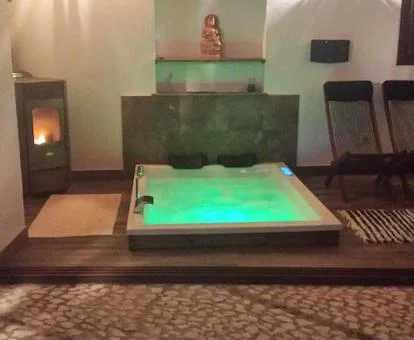 Jacuzzi a ras de suelo para dos personas con las luces verdes de hidroterapia y los chorros de hidromasaje activados, junto a dos sillas y la chimenea en el patio interior de la Casa Rural el Castaño