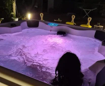 Enorme jacuzzi para cuatro personas que se encuentra en el exterior de la Villa Casa Prana donde vemos el mobiliario de la terraza y los árboles al fondo y una mujer relajándose en el interior del jacuzzi con la luz morada funcionando y los chorros de hidroterapia.