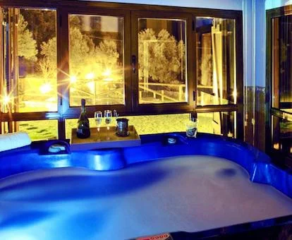 Foto del jacuzzi enorme para cuatro personas con una botella de cava y unas copas en el border y el exterior iluminado por la noche donde se ve el exterior nevado.