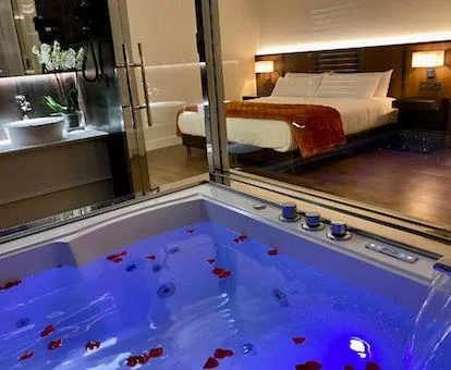 Foto del jacuzzi con luz azul y pétalos de rosa flotando sobre el agua y donde se puede ver la cama al fondo.