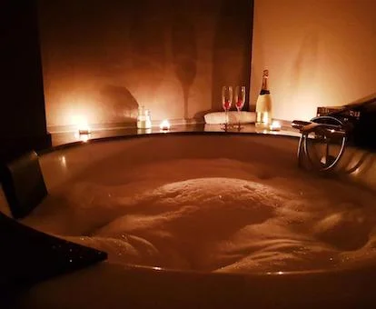 Foto del jacuzzi de forma circular para dos personas lleno de agua y espuma con dos copas de cava y una botella junto al borde en el apartamento deluxe con bañera de hidromasaje